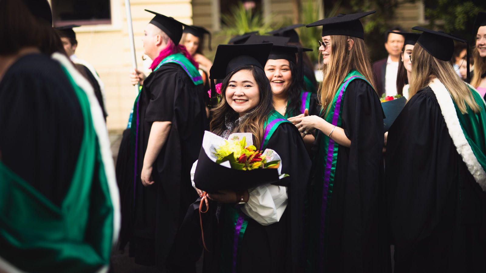 Graduates in caps and gowns gathered outdoors, one holding a bouquet.