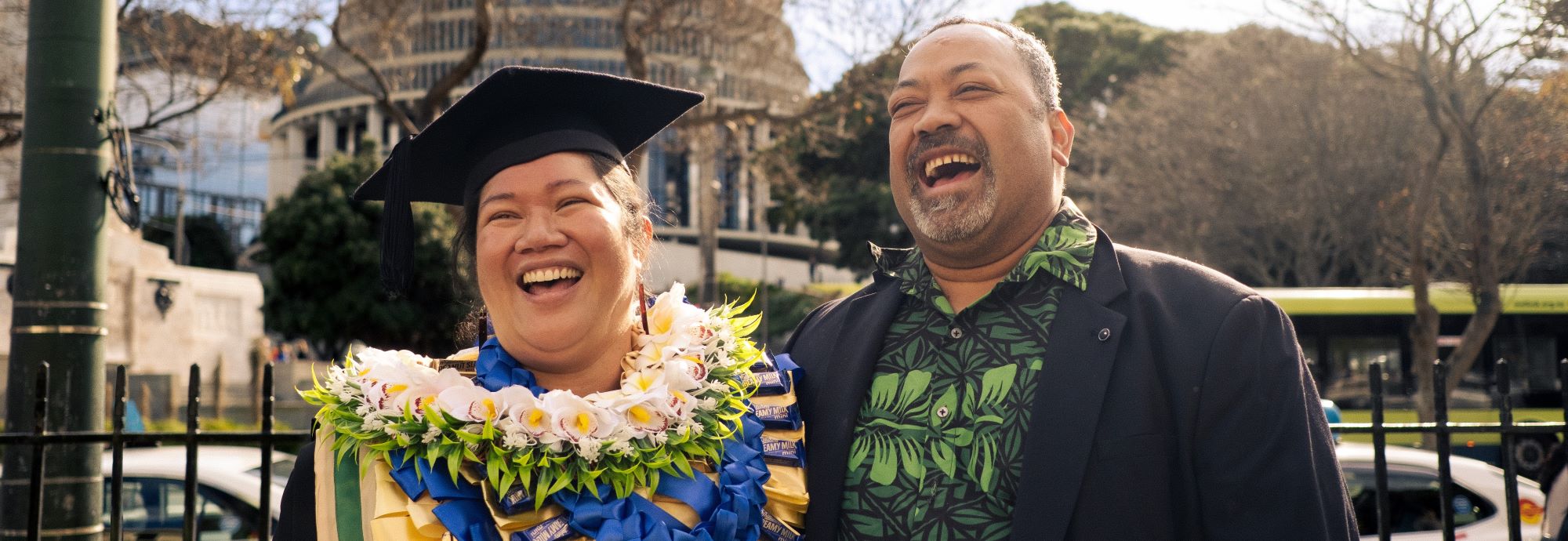 PG student wearing graduation robes, standing with a family member outside the Beehive