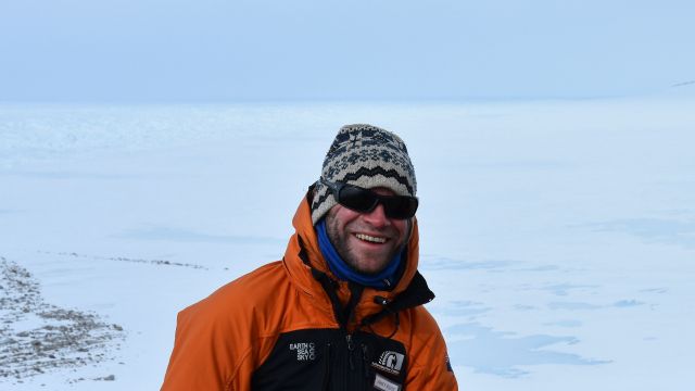An image of Jamey Stutz in Antarctica, he smiles, while wearing an orange jacket and cool sunglasses.