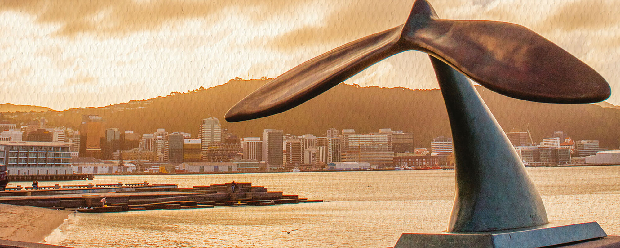 A two-tone image of a monument in the shape of a whale's tail, on a shore with Wellingon in the background.