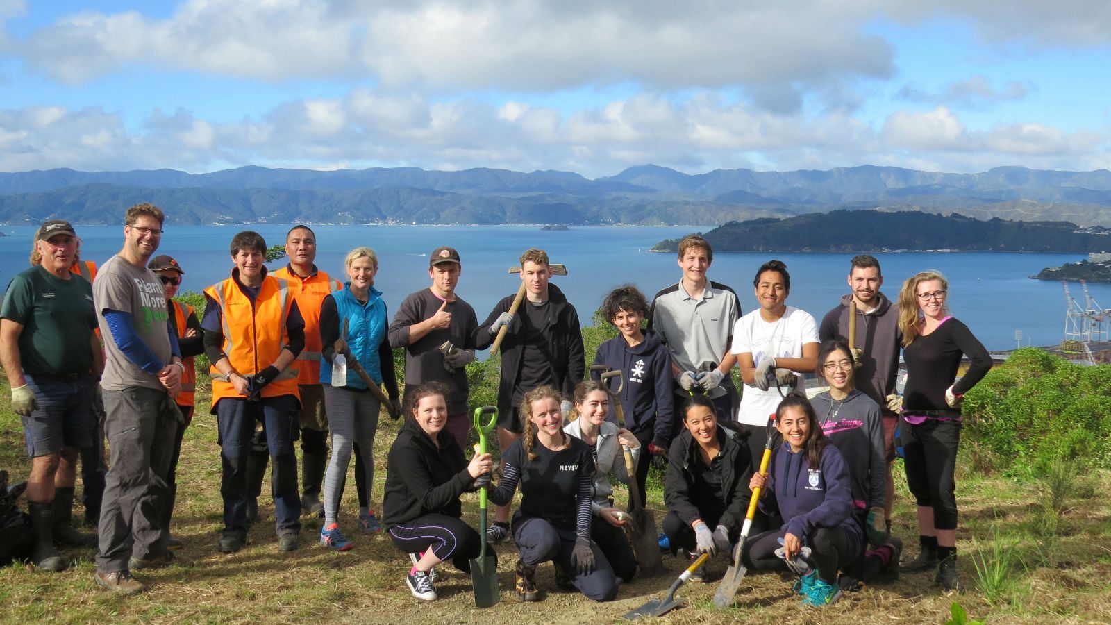 A group of student volunteers, hold landscaping tools while posing together on a hill with the ocean in the background.