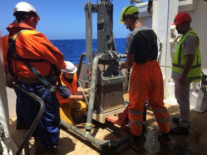 4 researches standing on a boat with one person kneeing in front of heavy machinery. Everyone is wearing safety clothing and it is a sunny day at sea. 