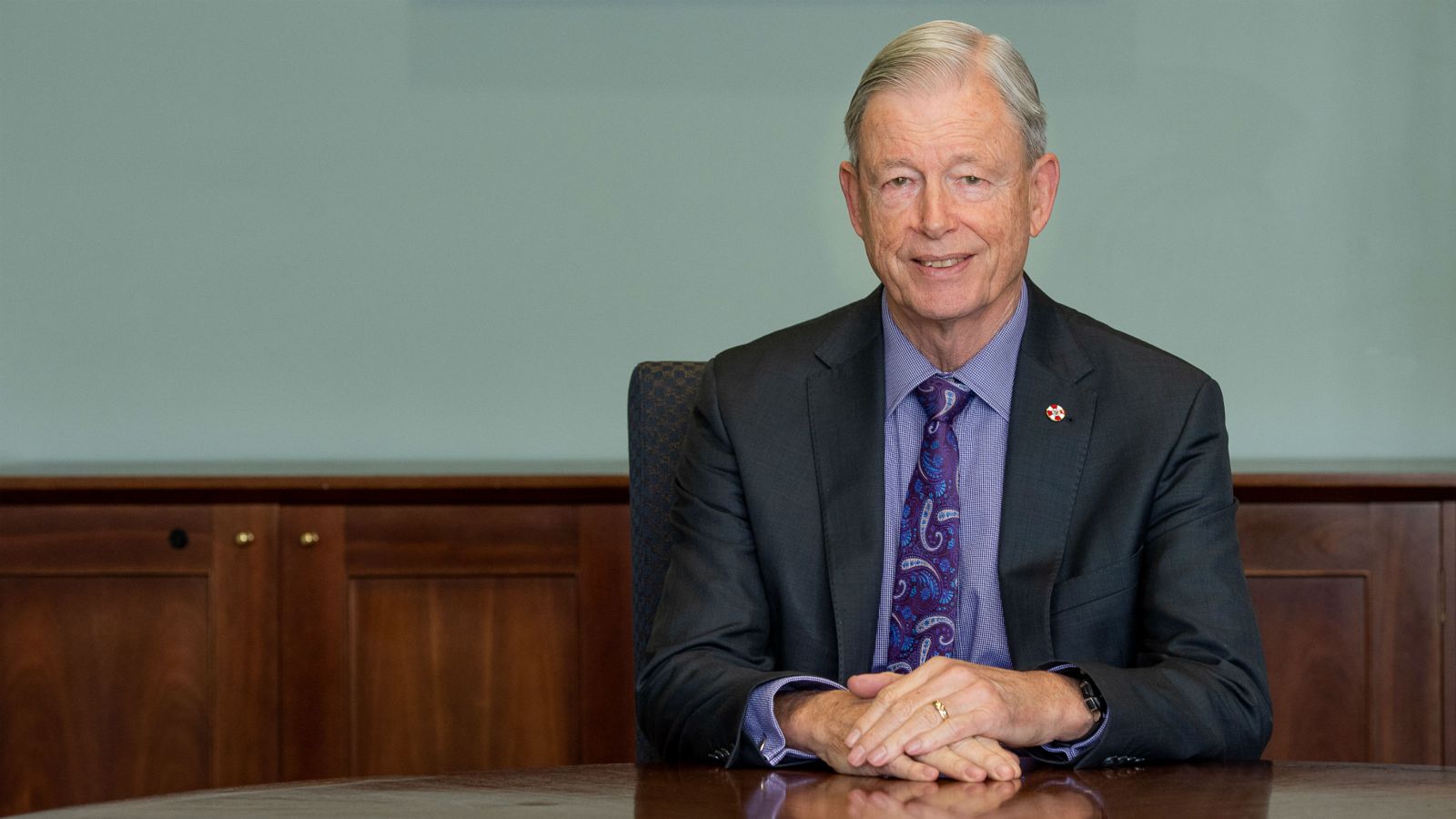 Professor Gregor Coster sits besuited between the dark, polished woodwork of a table and a wainscot wall.