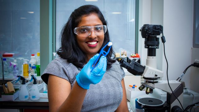 Dr Mima Kurian, wearing safety glasses and a blue glove, holds up a small piece of shell with a pair of tweezers. She is standing next to a microscope. There is a bench with lab equipment and windows in the background.