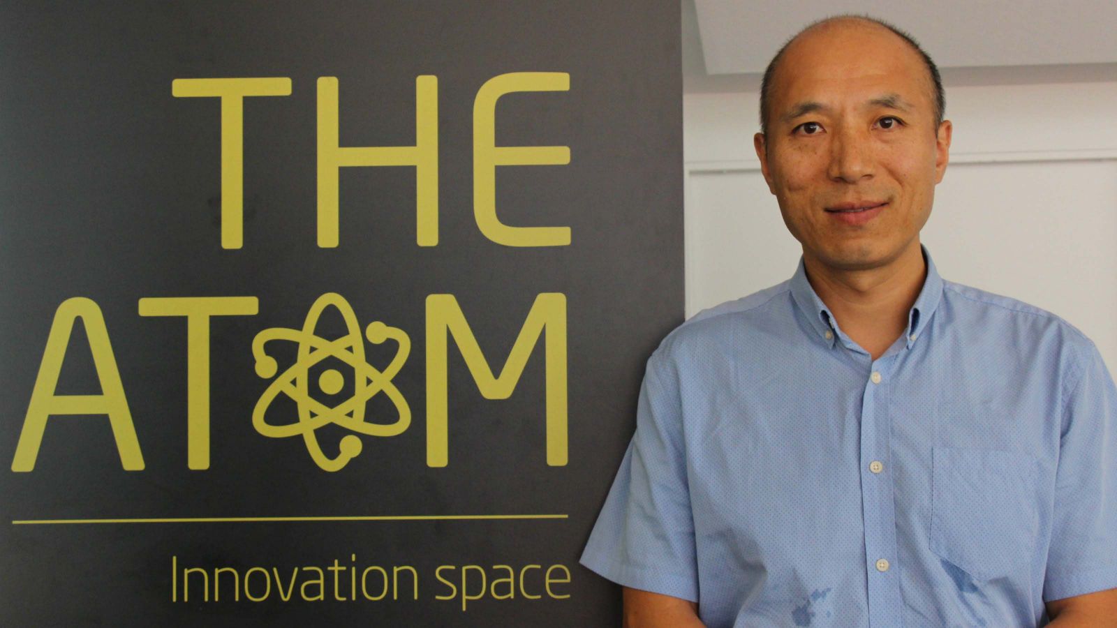 A profile shot of Hongzhi Gao standing by a banner that reads, The Atom.