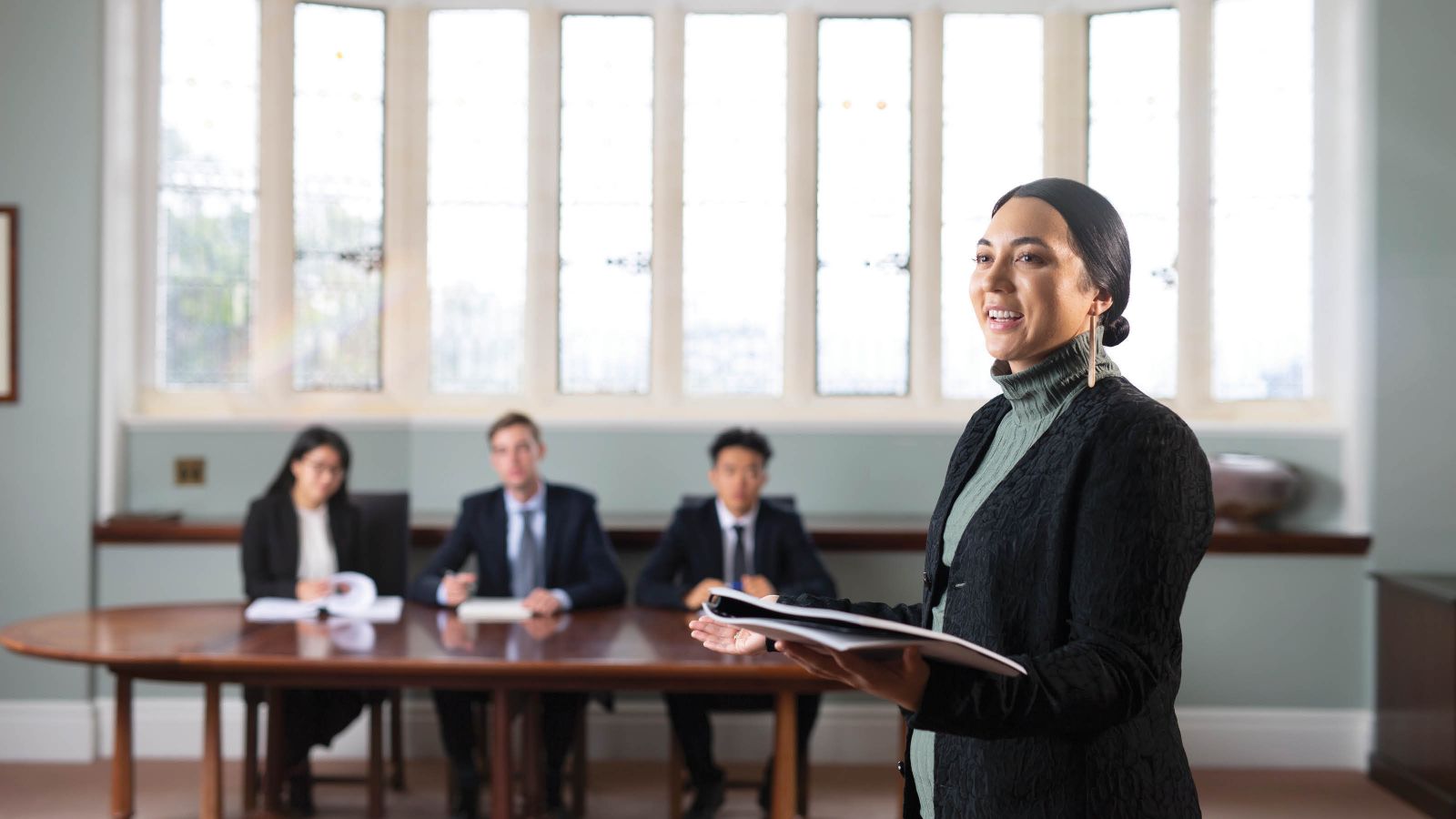 A female undergraduate law student standing, holding a document, and mooting in front of a panel of three judges who are seated at a table.