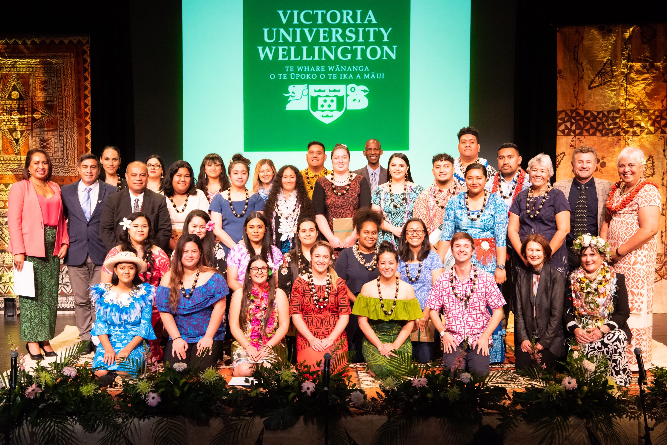 Pasifika graduation May 2019 – Four rows of people in colourful clothing smile together on a stage under a banner that reads Victoria University of Wellington.