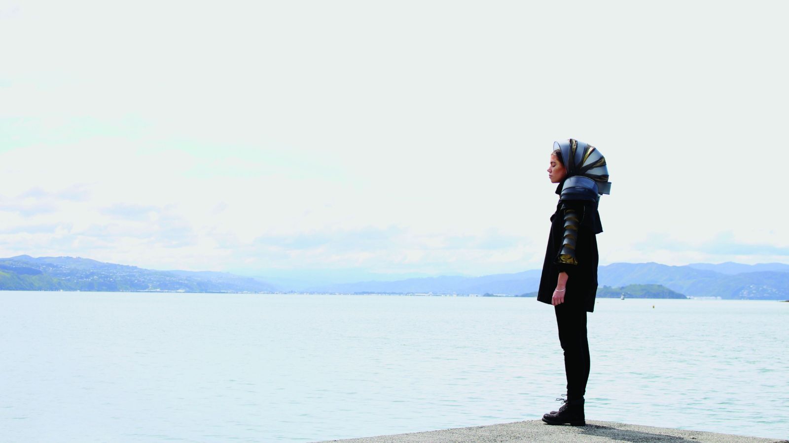 A woman in futuristic garb stands side on with Wellington Harbour in the background