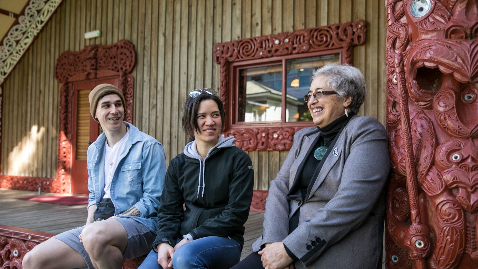 A boy, a girl, and an older woman sitting outside a marae chatting and laughing.