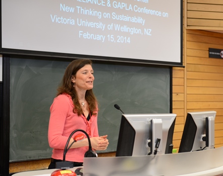 A professional woman wearing a pink shirt speaks at the front of a lecture hall with a screen in the background that reads, New Thinking on Sustainability.