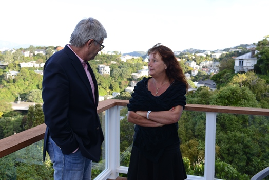 Two professionals converse on a balcony with lush green hills in the background.