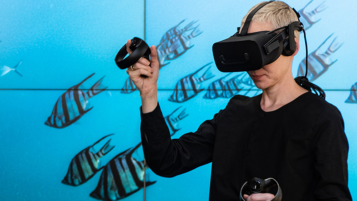 Woman wearing virtual reality headset and holding virtual reality handpiece, with background of fish swimming in blue sea.