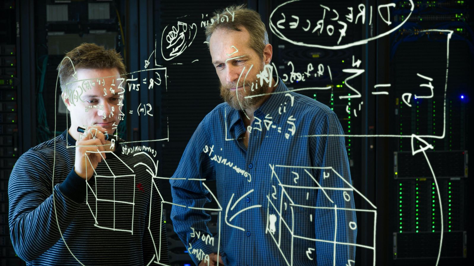 two scientists writing equations on a see-through white board