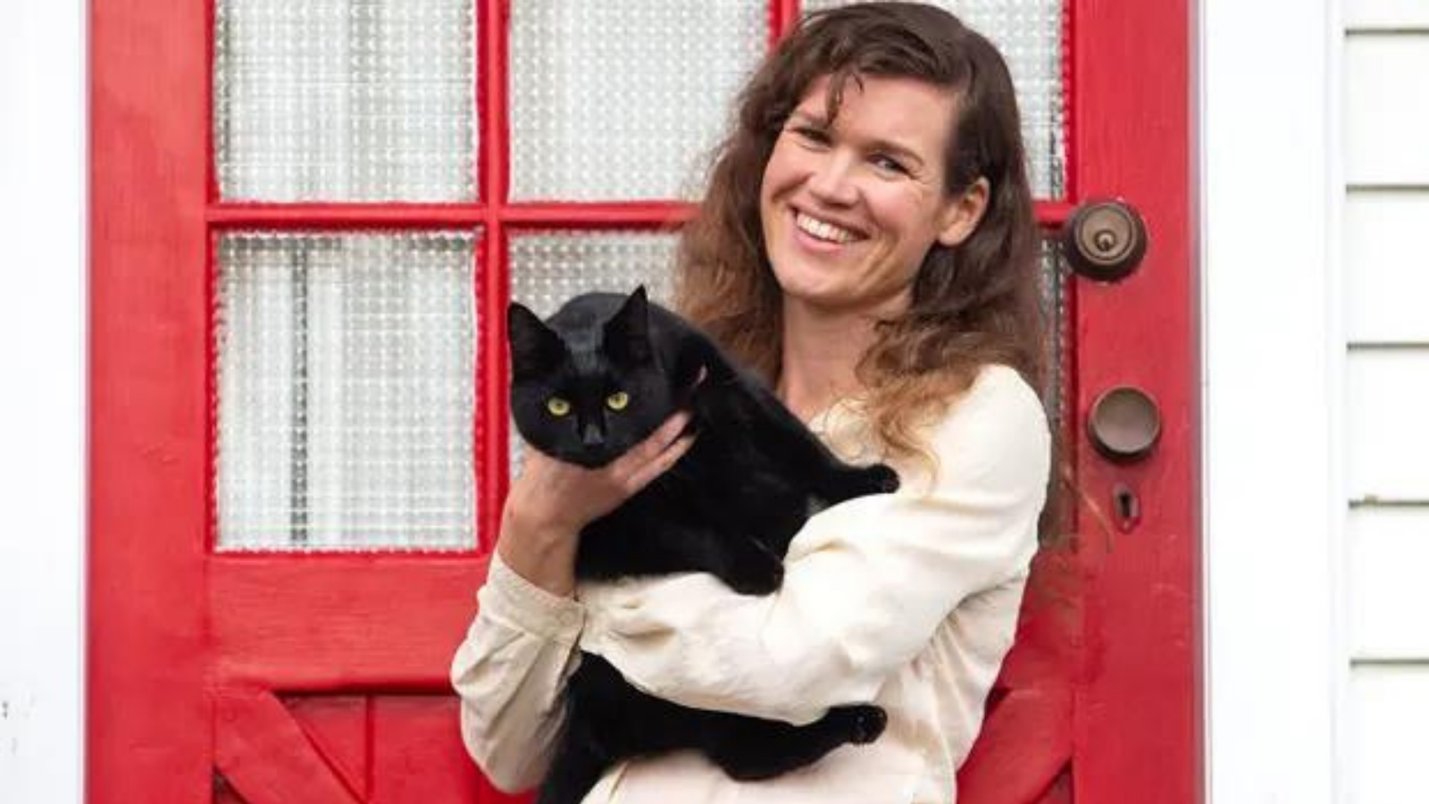woman holding black cat standing in front of red door with white house