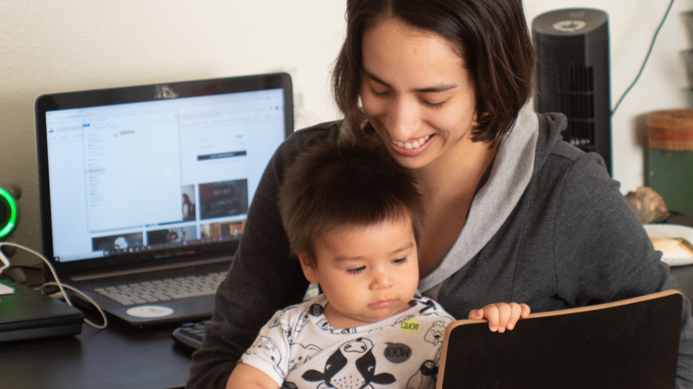 A young Asian woman holding a small child with a laptop computer on the desk behind her.