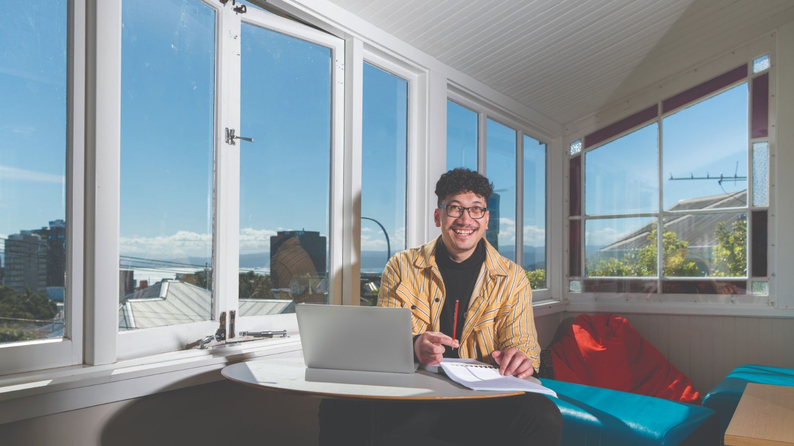 A smiling male student looks up from where he's studying in the Pasifka Haos, with a view of Wellington behind him.