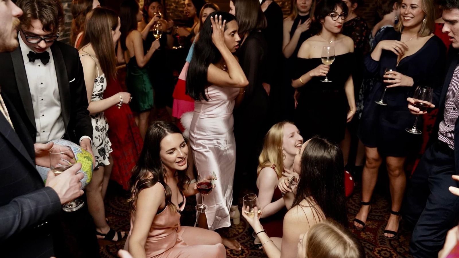 Students in formal dress, sitting, standing, dancing and drinking wine at French ball.