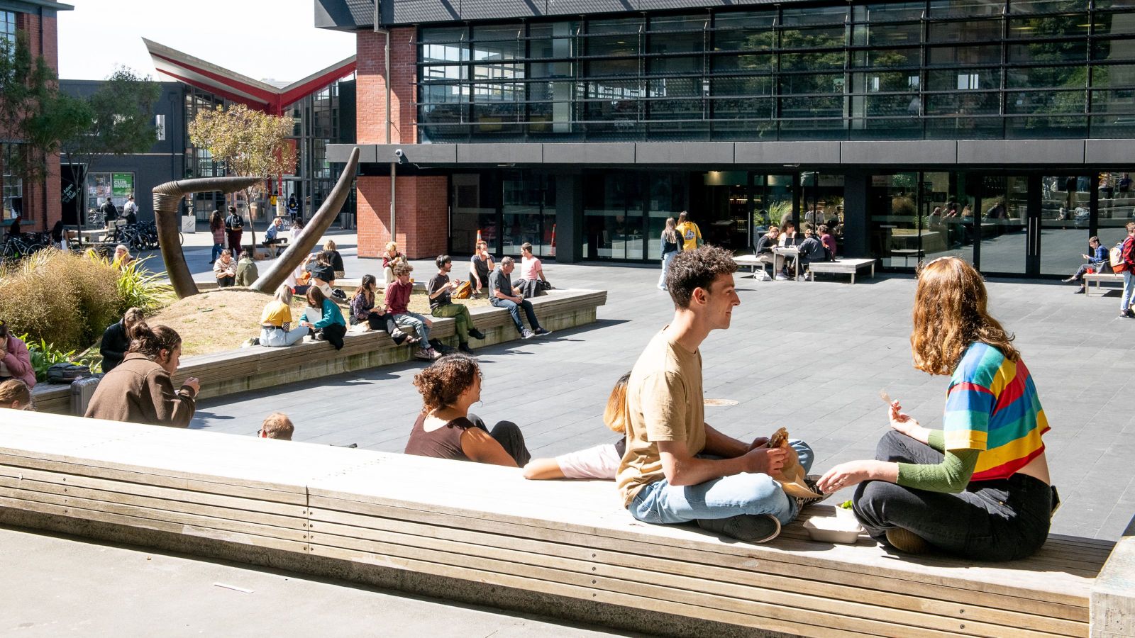 Beaglehold courtyard at Kelburn campus on a sunny day, with groups of students sitting, chatting, eating lunch.