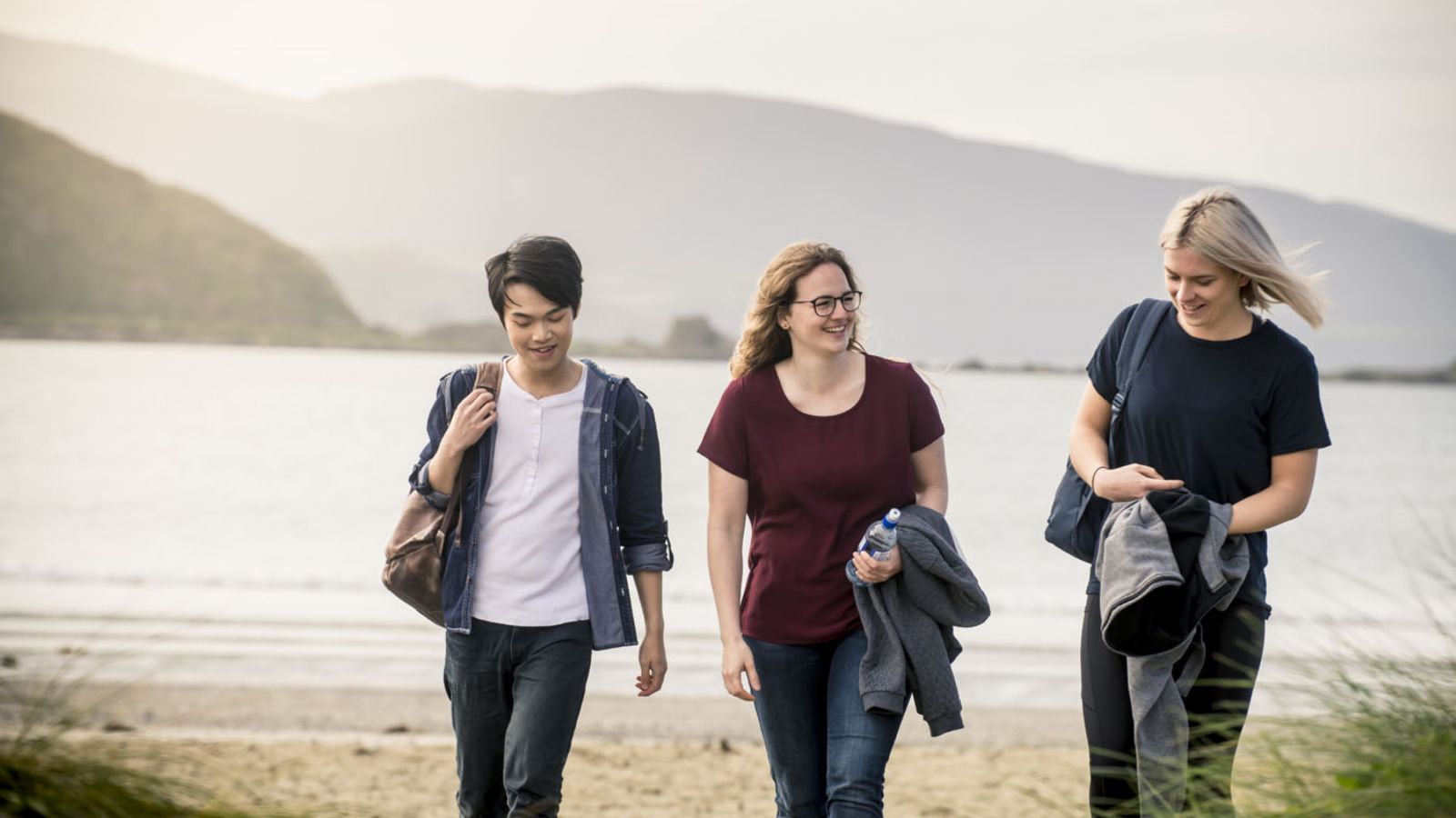 Three students walking across a sun-streaked beach in Lyall Bay