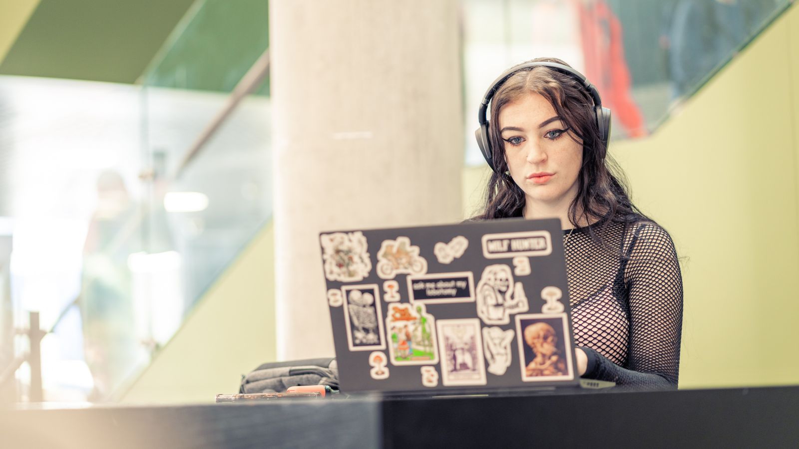 A female student wearing headphones and working on her laptop, which is covered in stickers, from a table in the Hub.