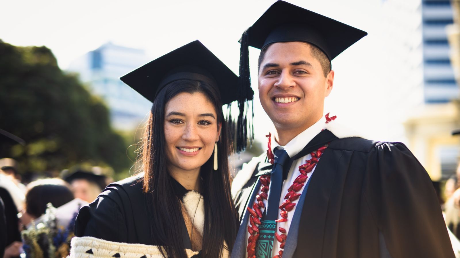 Two smiling graduates wearing academic gowns and caps pose together outdoors on graduation day.