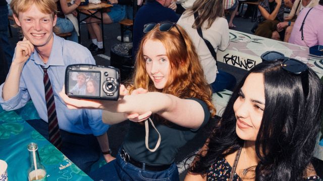 Three students at a street food market, sitting at a table taking selfies with a digital camera