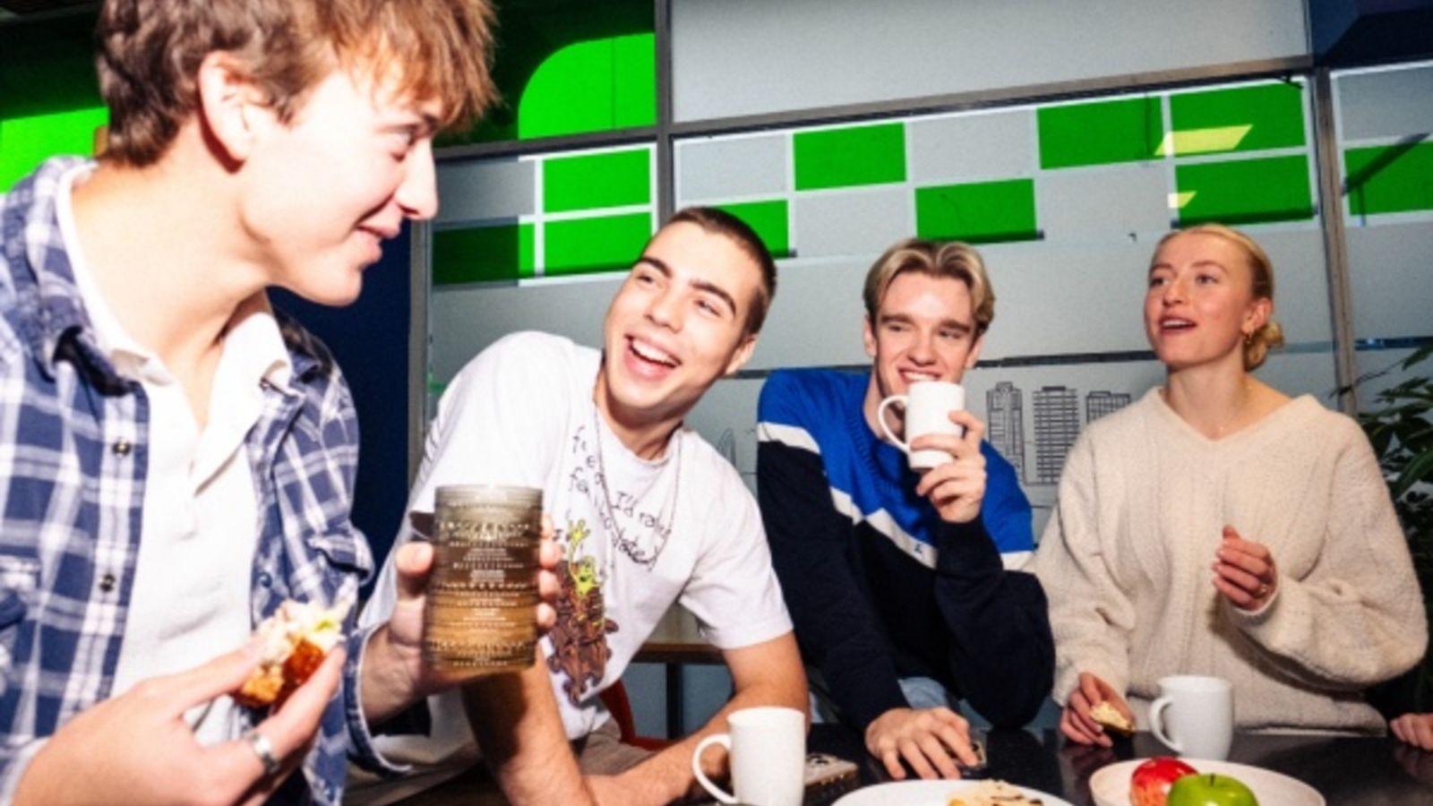 Four halls residents sitting around a table with food, drinks; a window with green and white patterns is in the background.