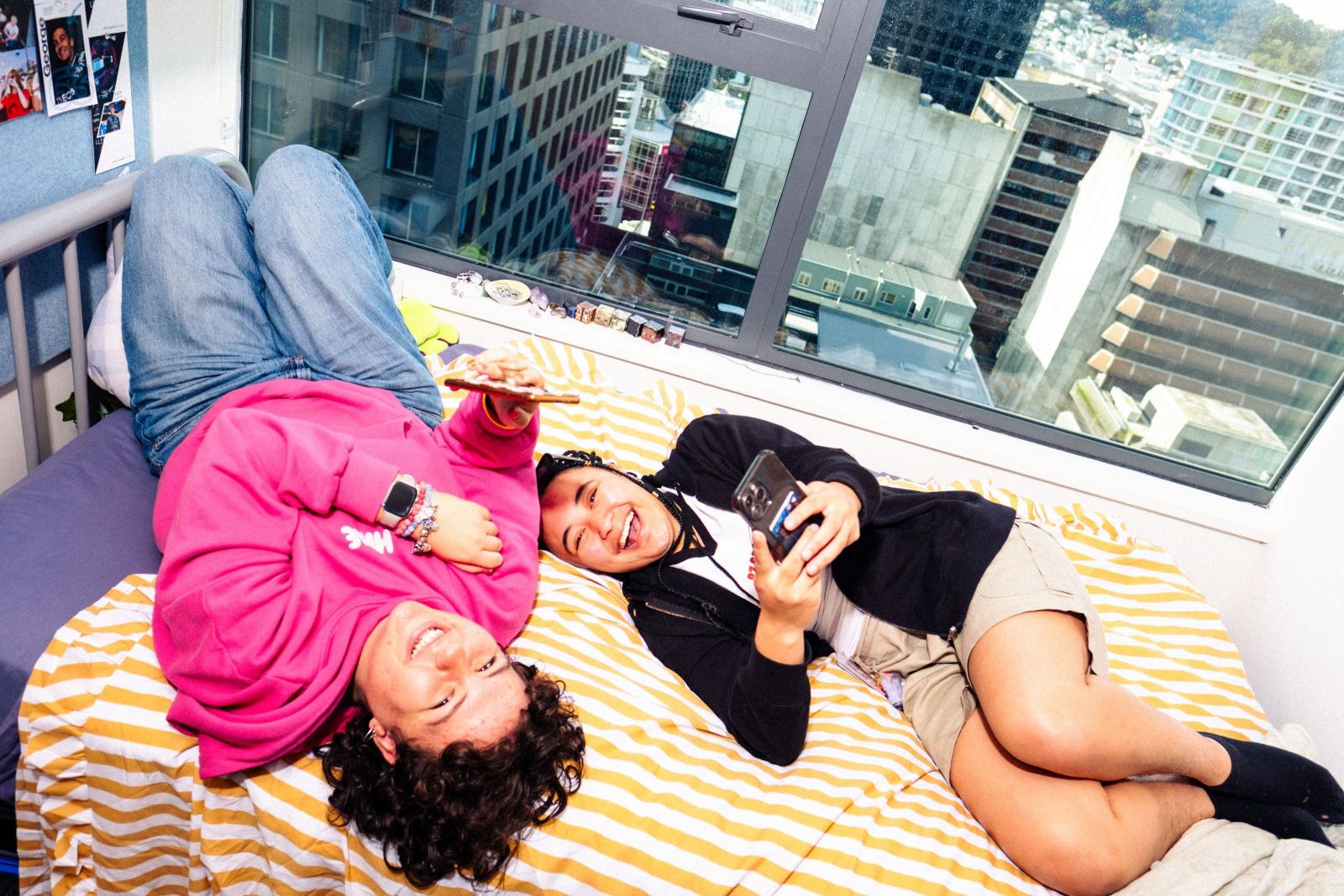 2 students lying on halls bed laughing with phones in their hands. Behind them is a window looking out to the city below