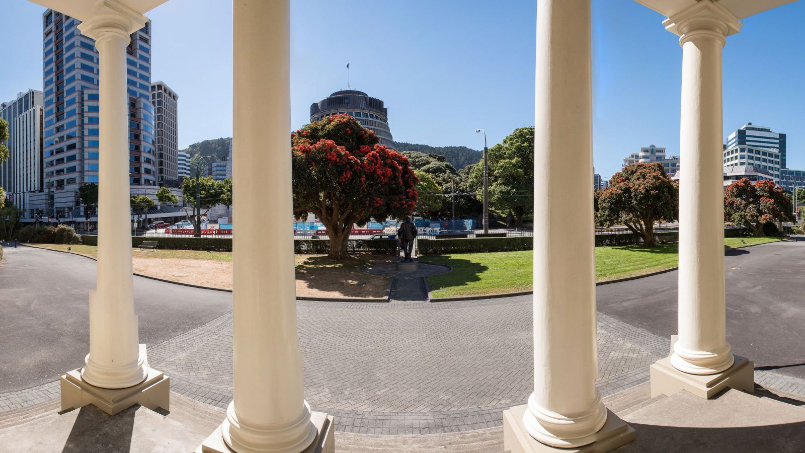 Panorama of Wellington’s Parliament precinct framed by white columns and pōhutukawa trees.