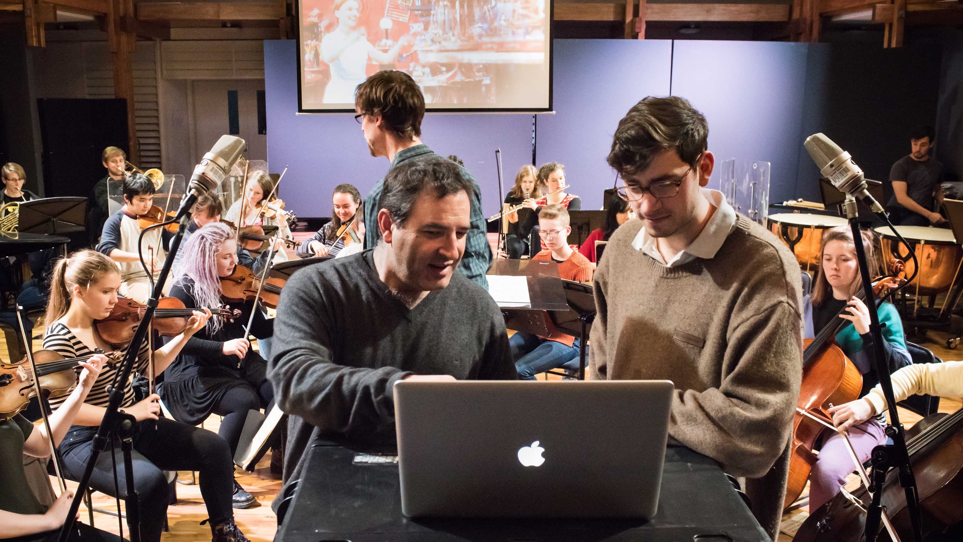 john Psathas and students – two men in the foreground look at a laptop while others play classical instruments in the background.