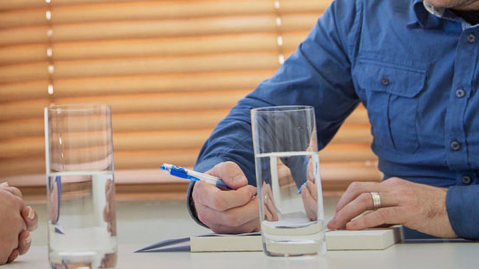 A man holds a pen to a pad of paper, on a table with two glasses of water.