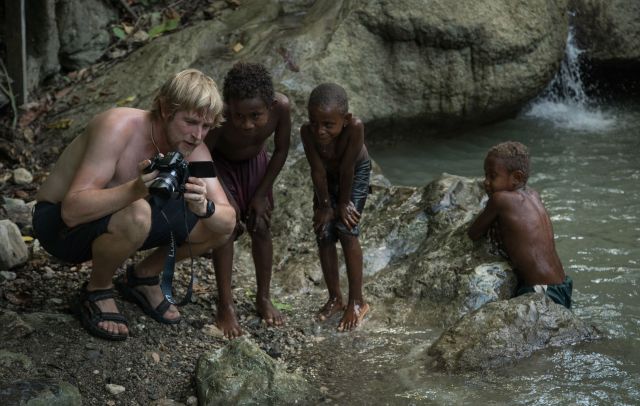 A man and some kids crouch by a stream looking at photos on his digital camera.
