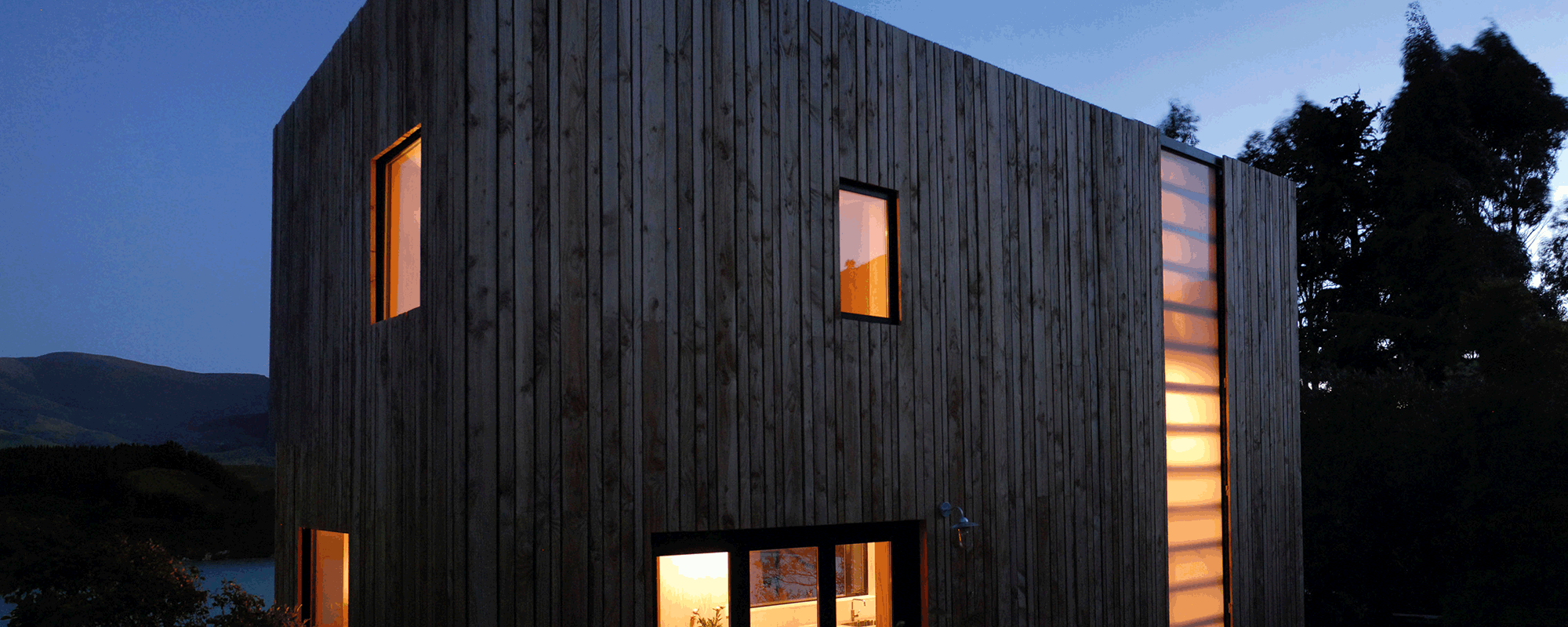 A small modern building, with vertical wood on the outisde taken at dusk, with warmly lit windows.