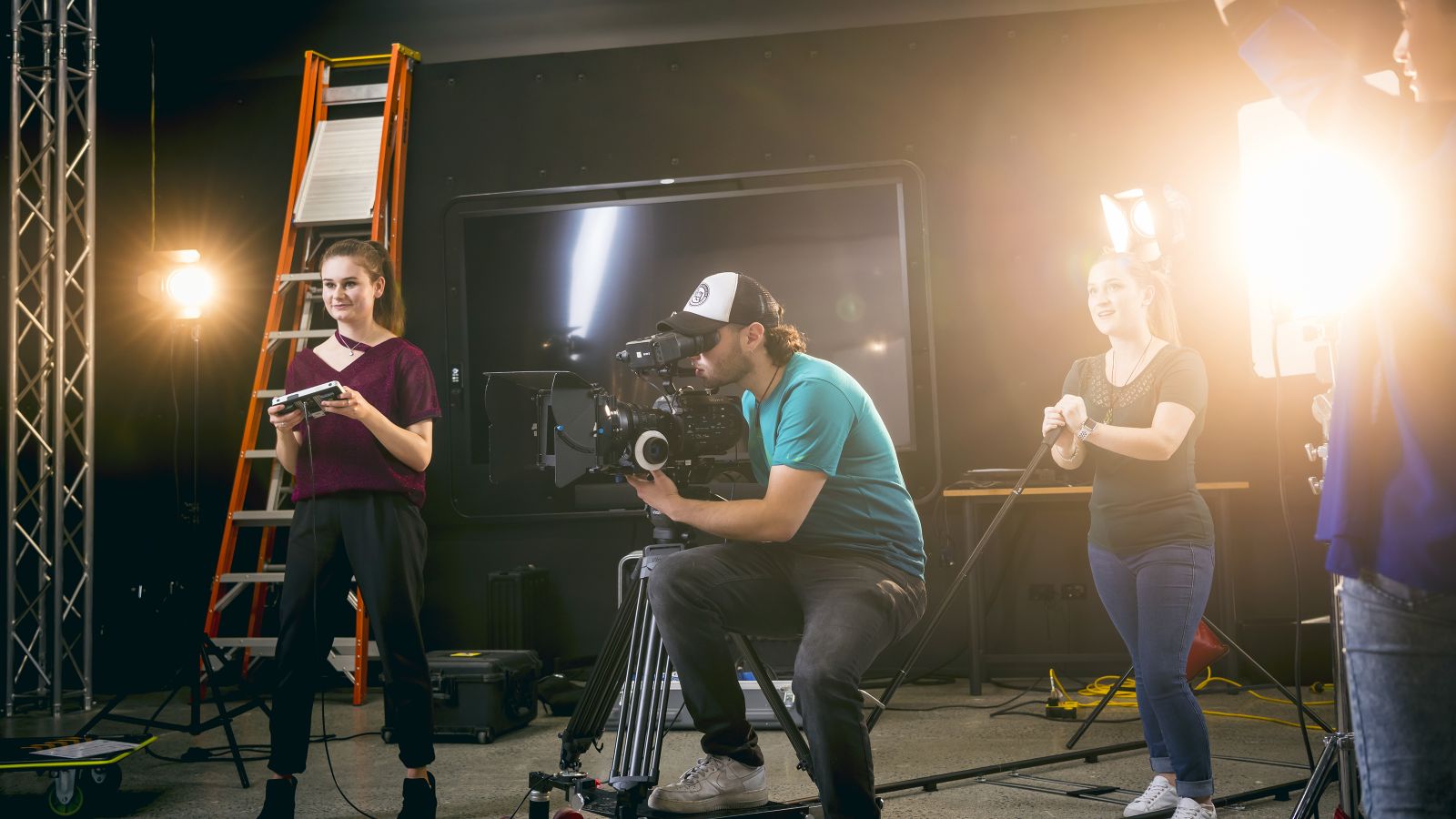 Students at work in a film studio, with a young man operating a camera in the middle and two females either side using other equipment.