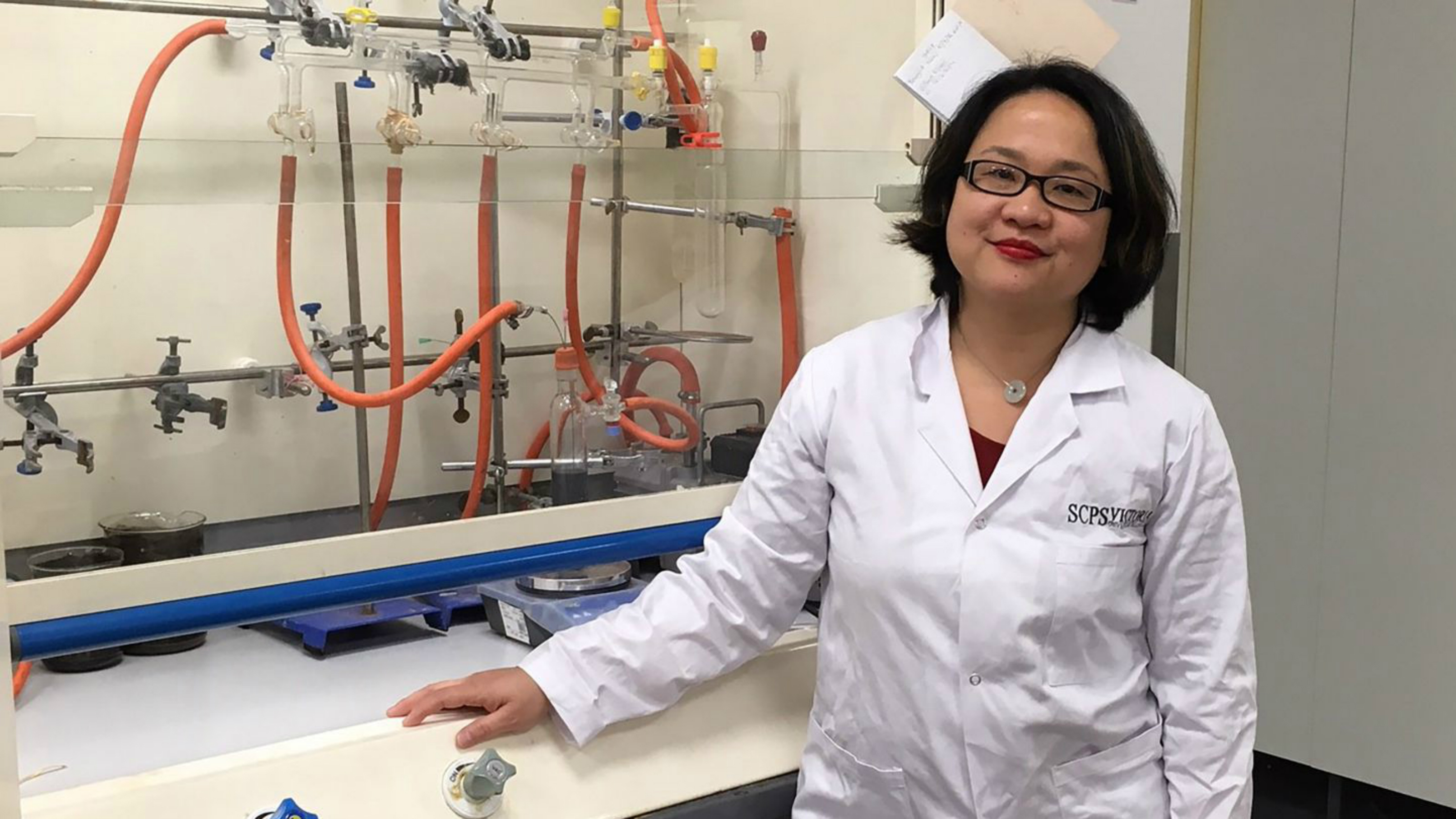 PhD student Putri Fraser, wearing a lab coat, stands by a lab set up.