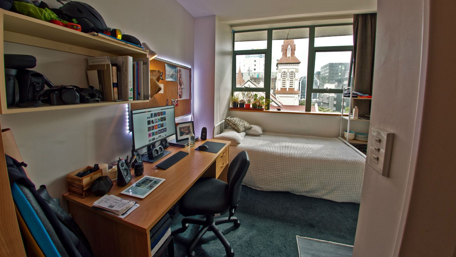 Single studio bedroom in Willis St – Education House. A room with a double bed, a desk and a book shelf. The view through the window overlooks the city.