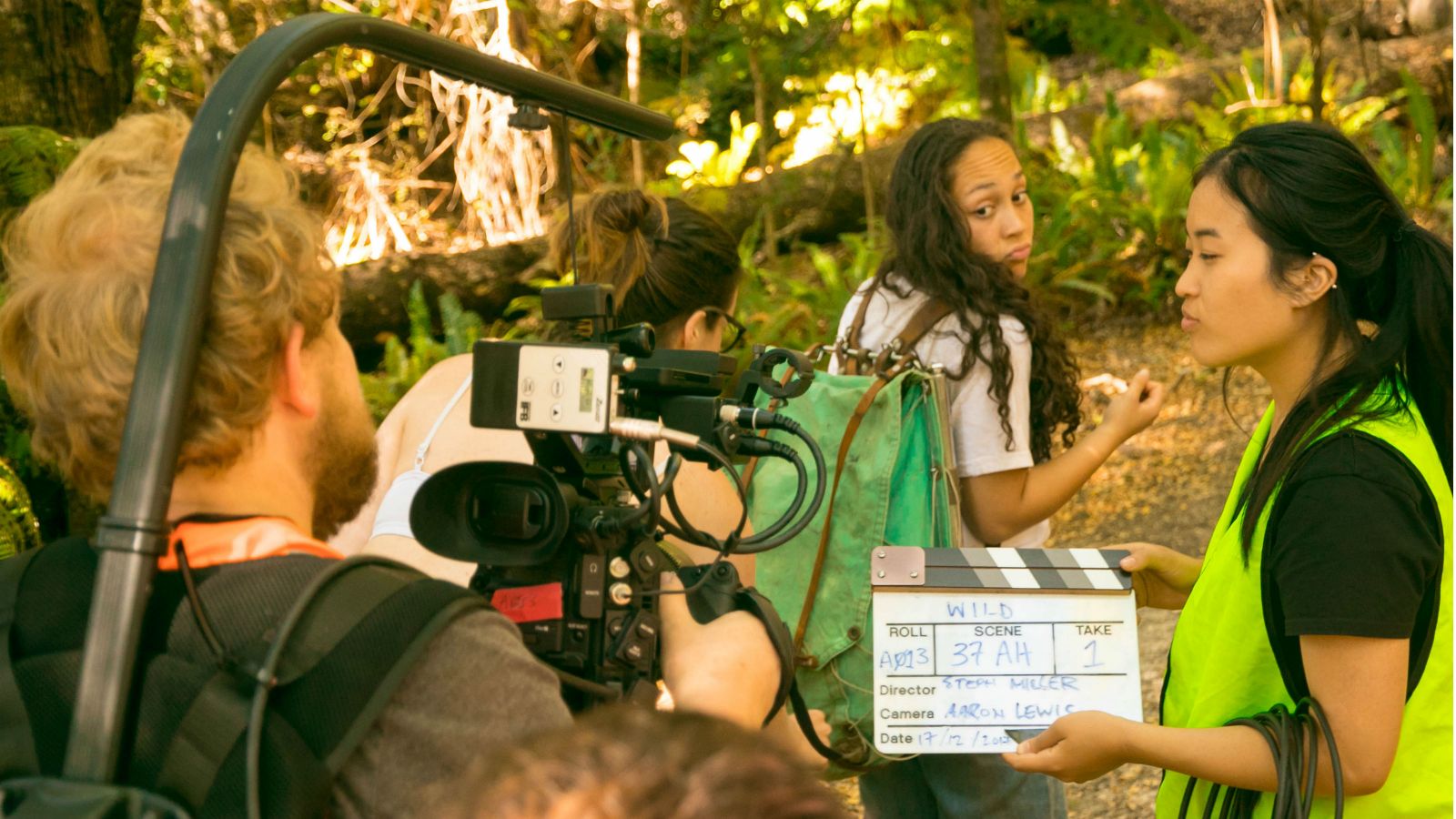 Students as cast and crew in a chiefly reddish yellow and green forest producing a film with essential filmmaking equipment, such as a video camera, clapperboard, cables, and highlighter vests.