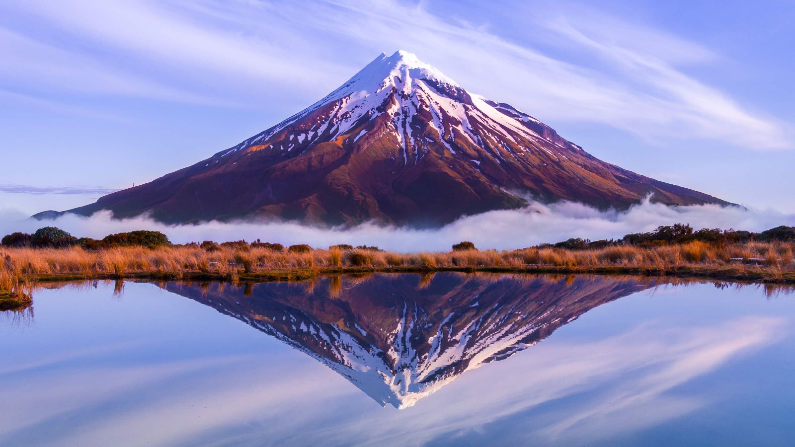 The symmetrical Mount Taranaki with its single, snowy peak extending into a soft violet sky wisped with white clouds. The entire monumental scene is perfectly reflected in the mirror of the lake separated from the mountain’s base by hot orange bush and a strip of low-lying cloud.