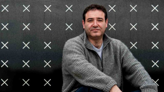 John Psathas sits in front of a wall with many x's on it.