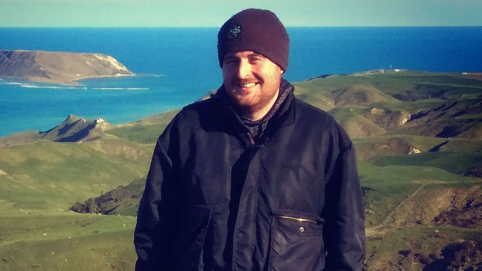 Harry, wearing a black jacket and beanie, stands in front of green hills and blue ocean. A small island can be seen in the distance. 