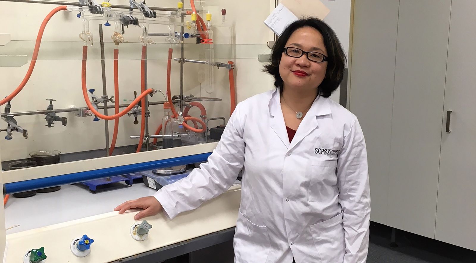 Putri, wearing a lab coat, stands next to chemistry equipment.
