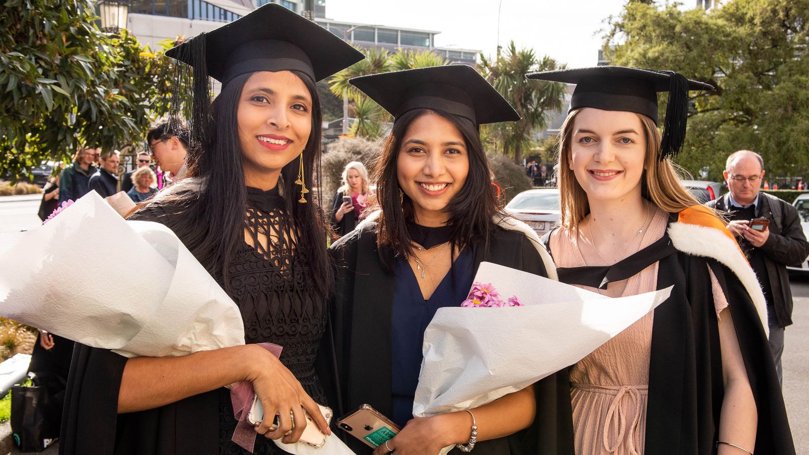 Three graduates, two who are holding flowers, smile for a photo during the graduate street parade.