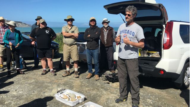 James talks to a crowd of people by a car. There are fossils on the ground. It's a sunny day and the blue sky and sea can be seen behind everyone. 