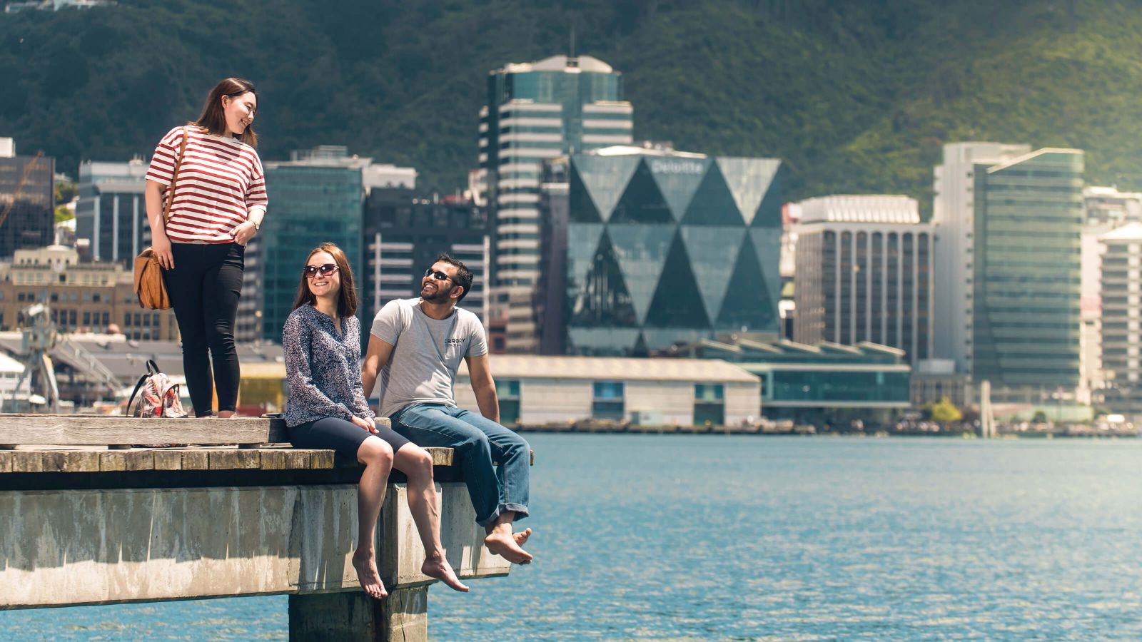 Three smiling students are standing and sitting on the edge of a wharf on Wellington's waterfront.