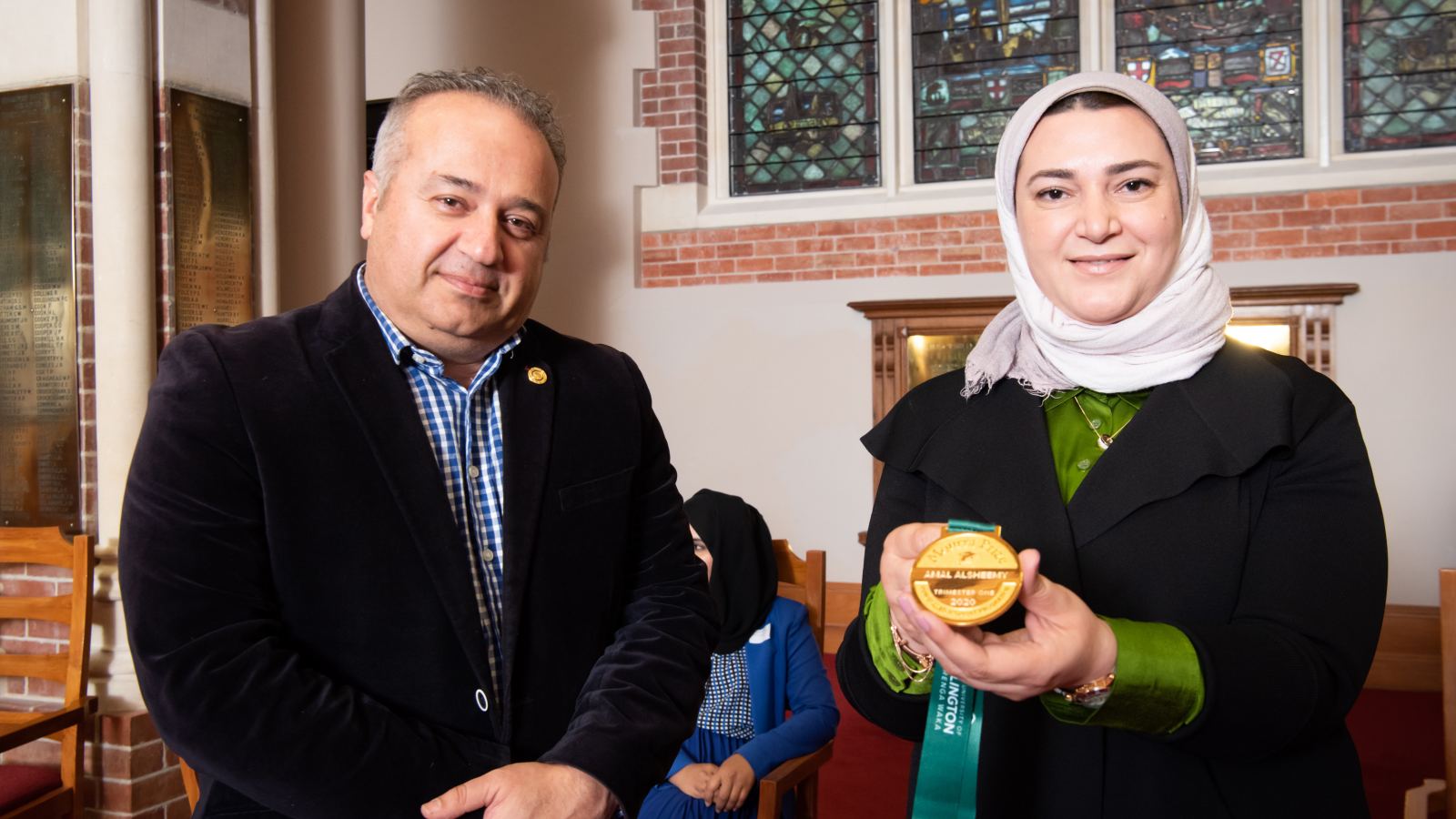 Man with suit and girl with pink headscarf showing medal in her hand, with stained glass window background