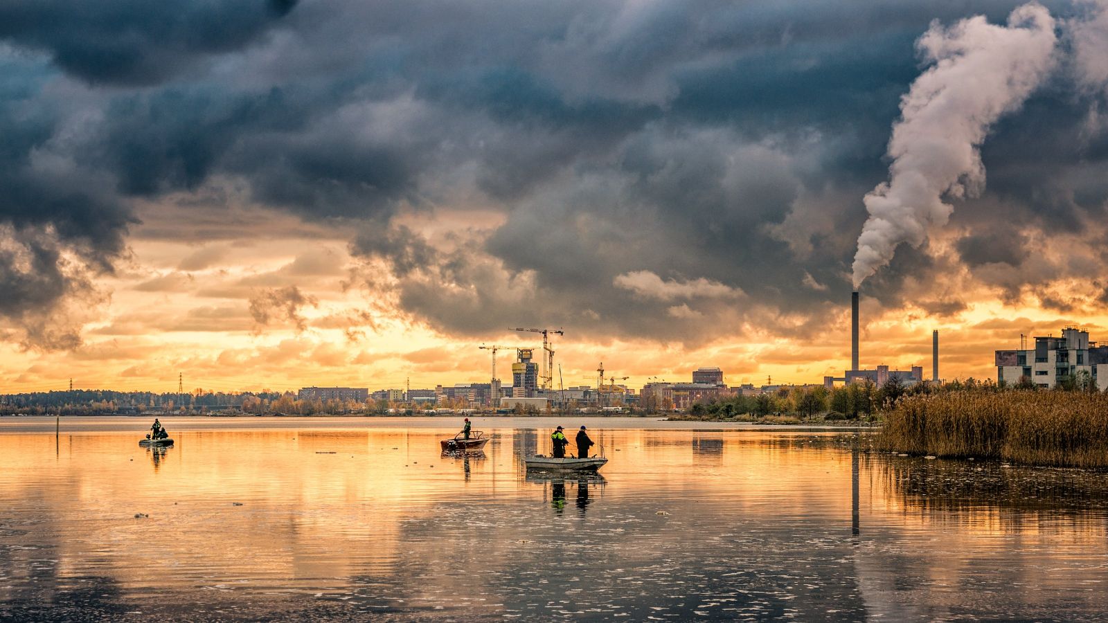 Mirror-smooth lake at sunset with small boats in foreground and dark clouds and a prominent smokestack in the background