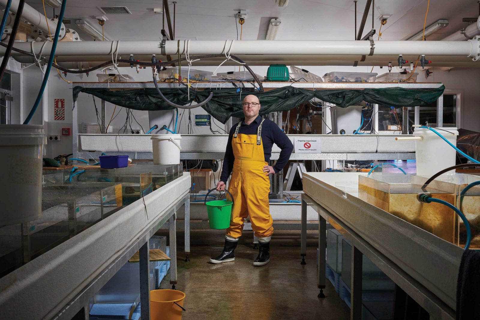 Dr Chris Cornwall standing in a marine lab wearing orange overalls and a green bucket. 