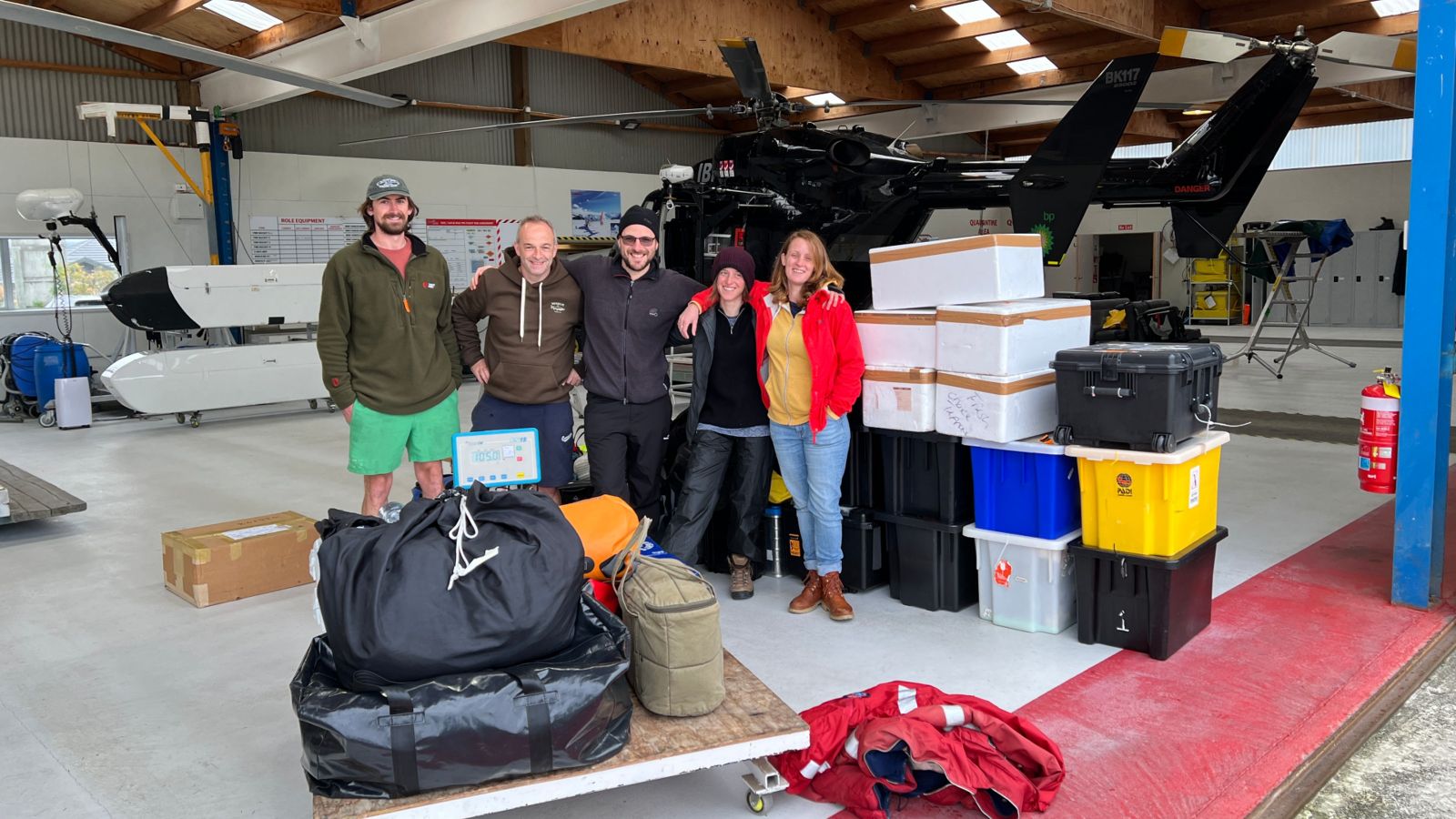 Research team with gear packed, waiting in helicopter hangar with helicopter in background.