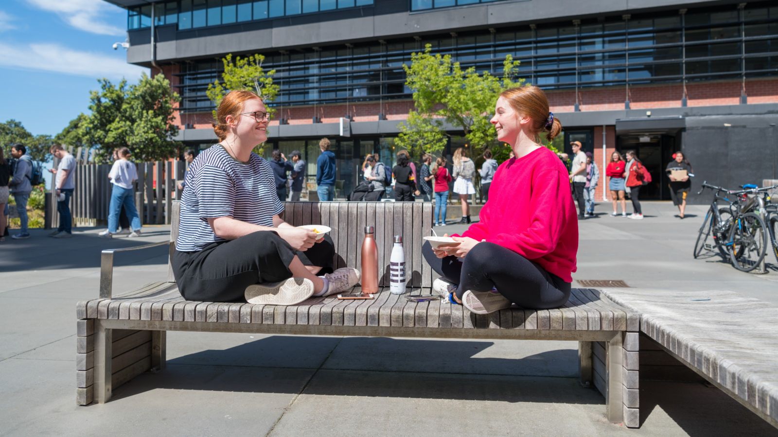 Two students sitting cross-legged on a bench facing each other and talking.