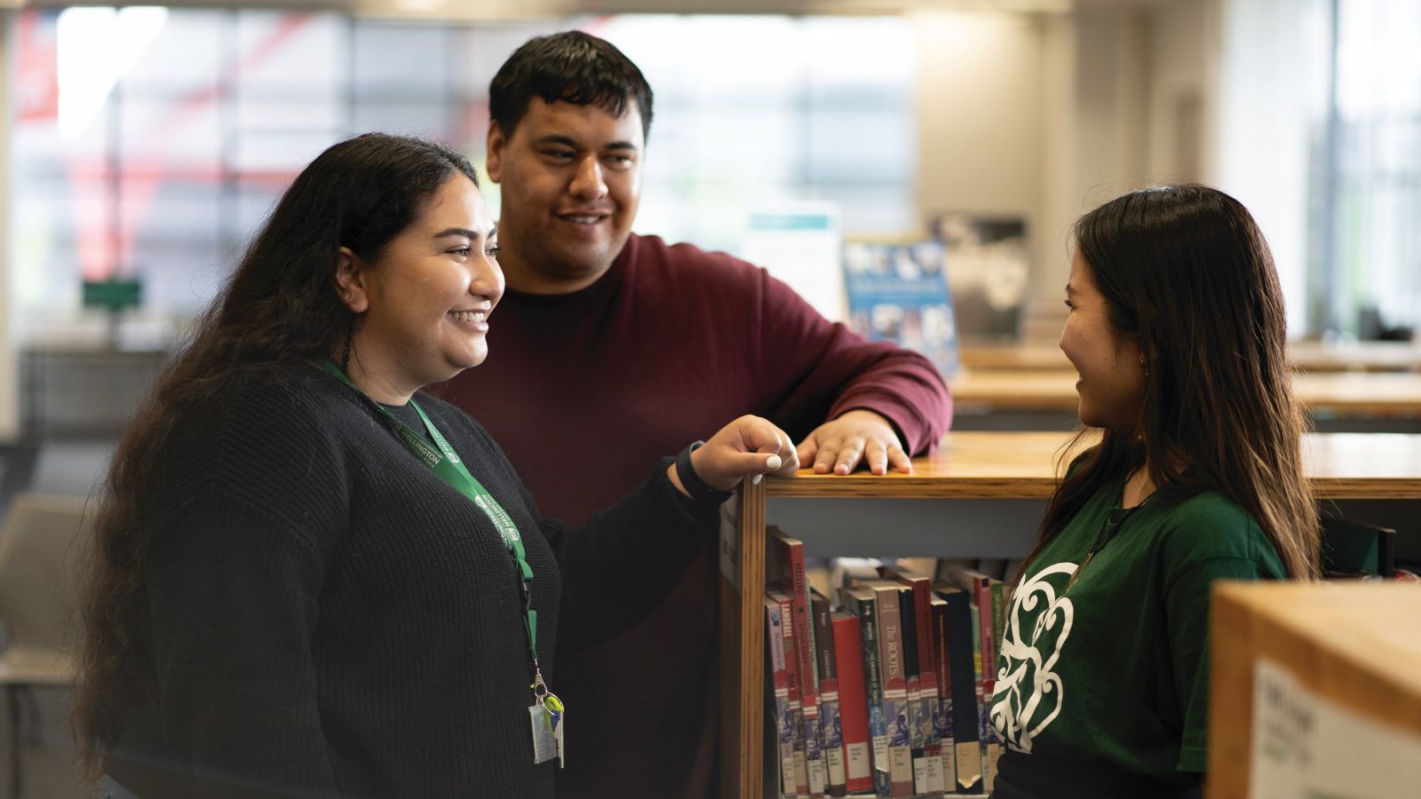 A young Māori woman and man talking to another student in front of a bookshelf.